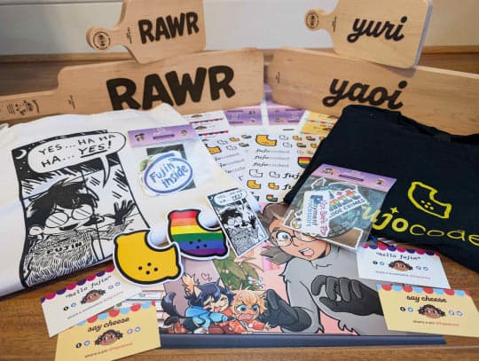 A variegated series of merchandise, from back to front: wooden cheese boards engraved with RAWR, yuri and yaoi, a tote bag featuring a parody of the YES... HA HA HA YES! Sickos joke (reading Fujin instead), a T-shirt with the fujocoded logo (a lemon), various stickers, a print and fujocoded business cards.