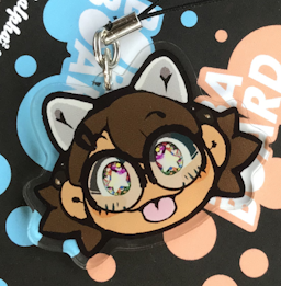 The Boba-tan blep charm photographed on orange and electric blue BobaBoard logos